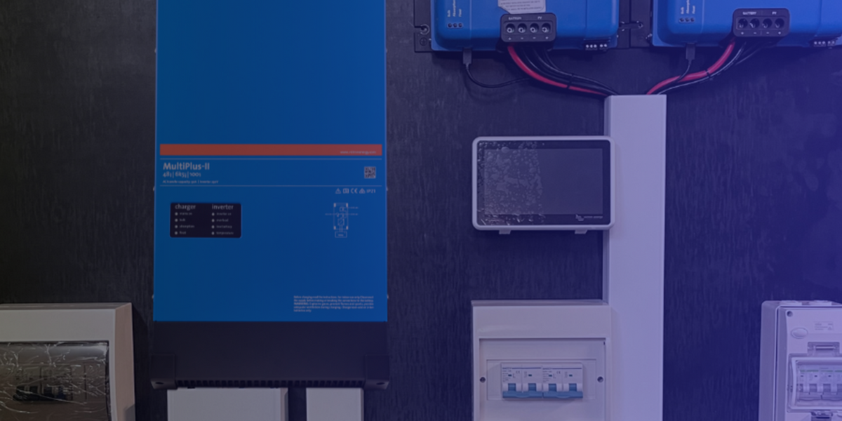 A large blue Victron MultiPlus 6 inverter panel mounted on a dark wall, alongside other electrical components including a small screen, circuit breakers, and blue battery units at the top.